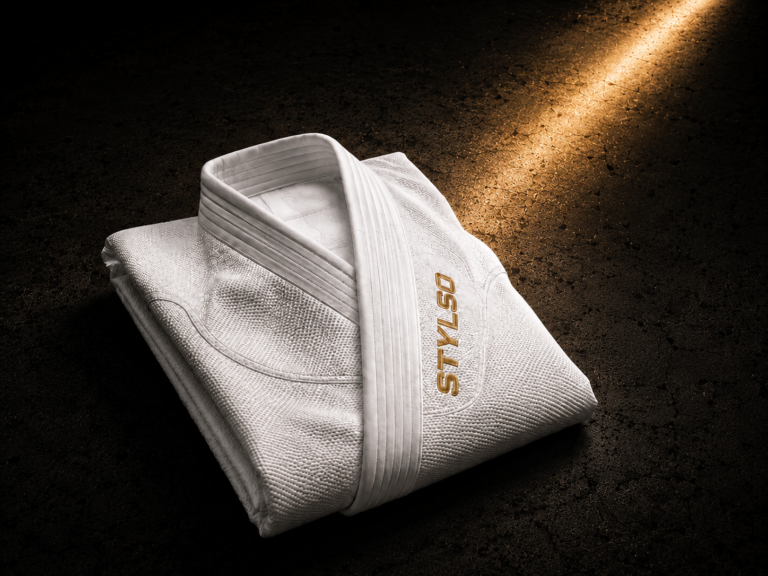 Stylso white pearl-weave BJJ gi laid flat on dark slate, gold accent lighting — classic competition colour for BJJ and judo